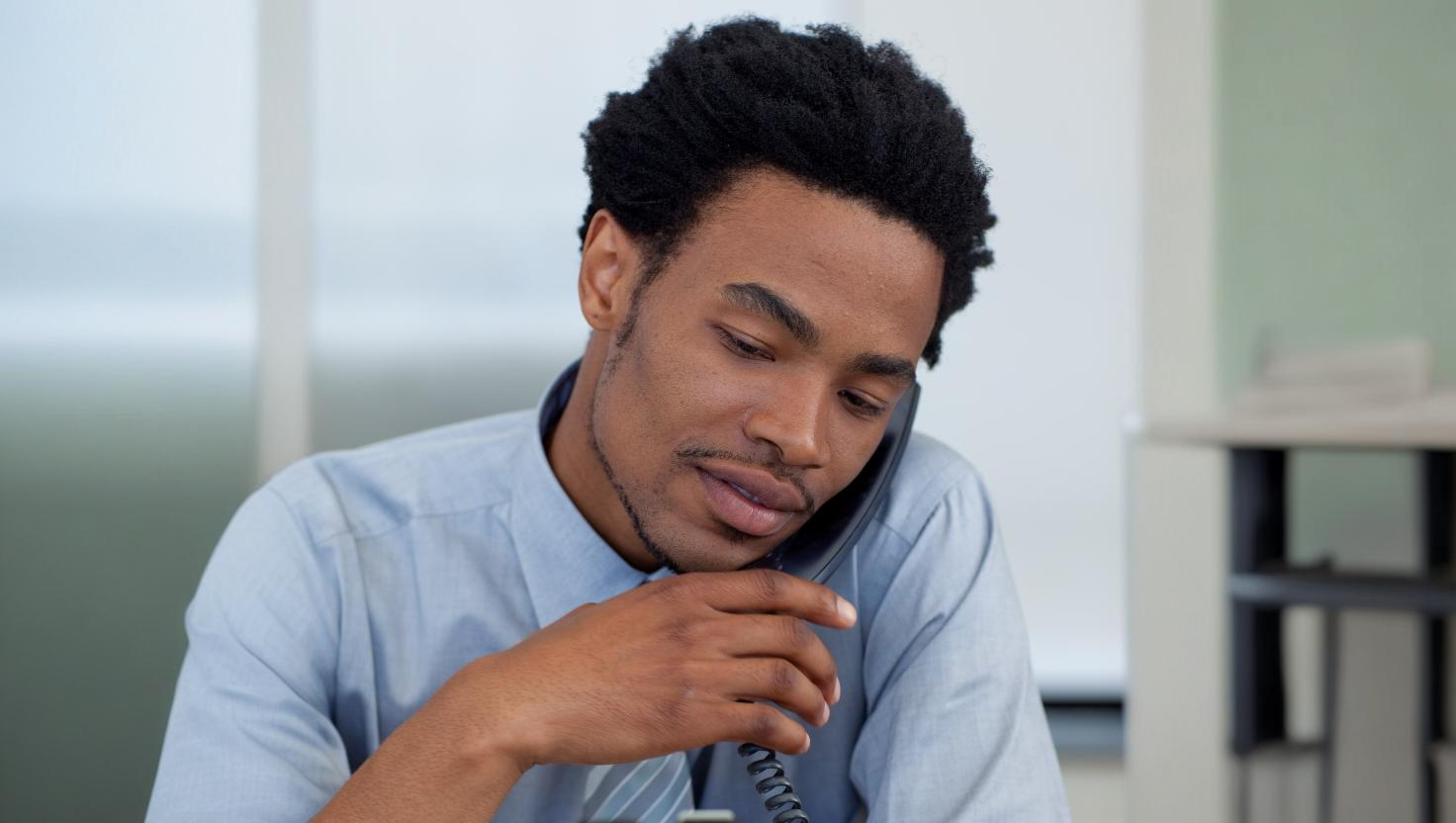 Man in a light blue shirt speaking into a corded desk phone, symbolizing a business call setup where Teams PSTN call recording could be implemented for communication monitoring or quality assurance.